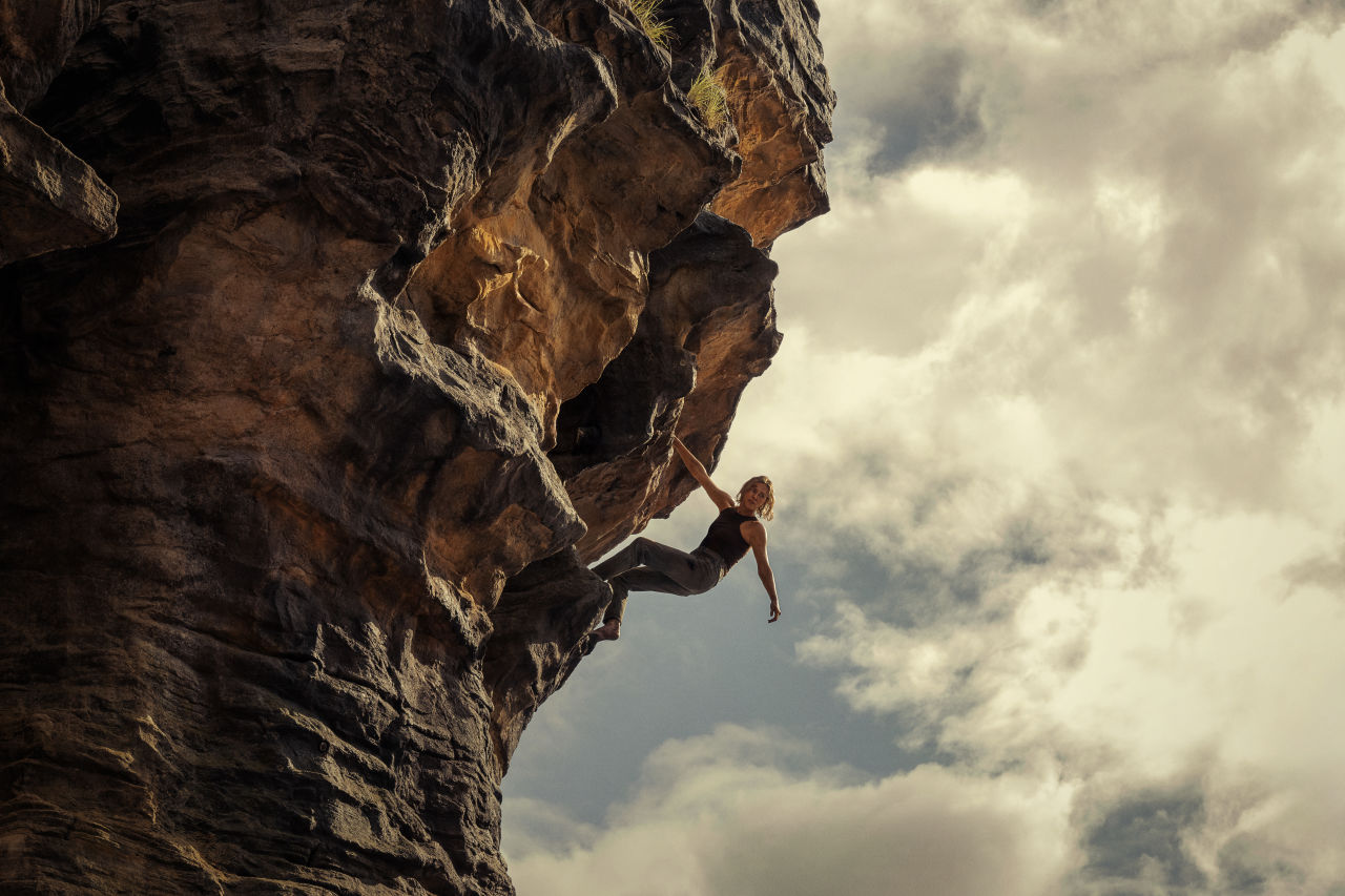 Charlize Theron as Sasha clings to the underside of a dramatic sandstone overhang against a cloudy sky in a scene from Netflix's Apex, filmed in the Blue Mountains of New South Wales, Australia.